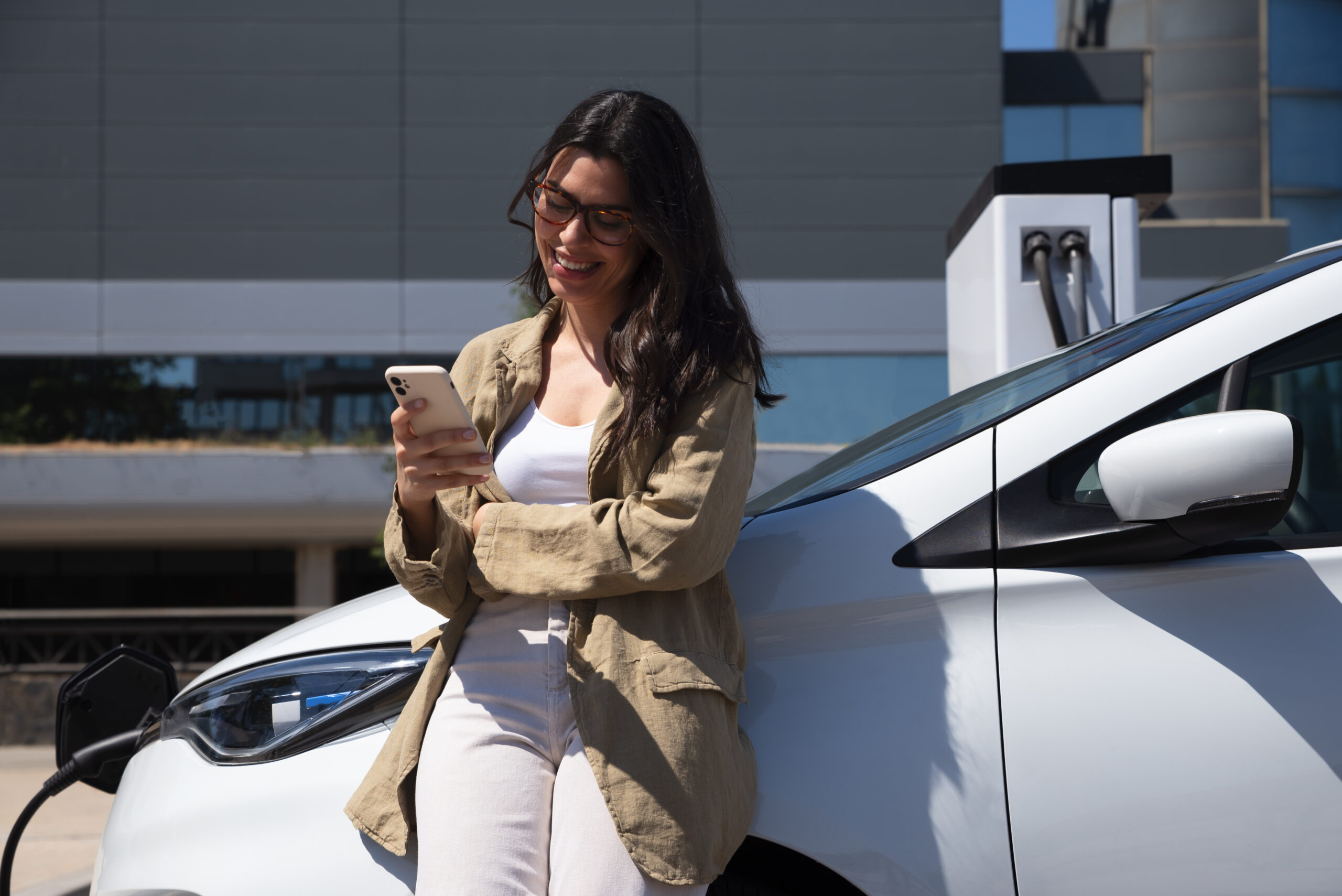 Mulher sorrindo enquanto usa o celular, encostada em carro elétrico durante recarga, representando mobilidade sustentável e novidades da CNH Digital 2025.